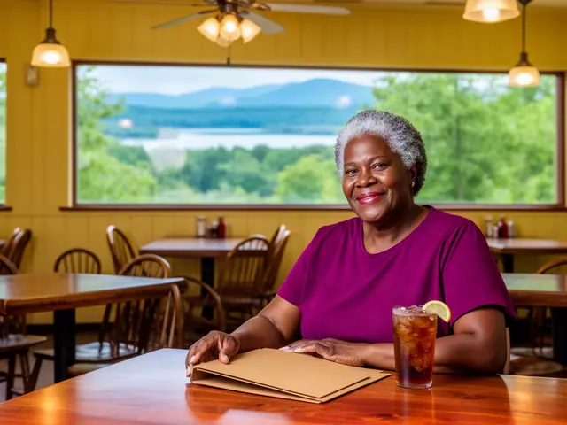 A smiling restaurant owner seated with a thin folder and iced tea in a sunny dining room with a lake view.